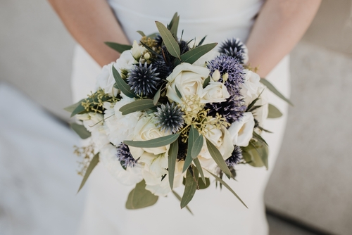 A bride stands holding a beautiful bouquet of roses and thistles.